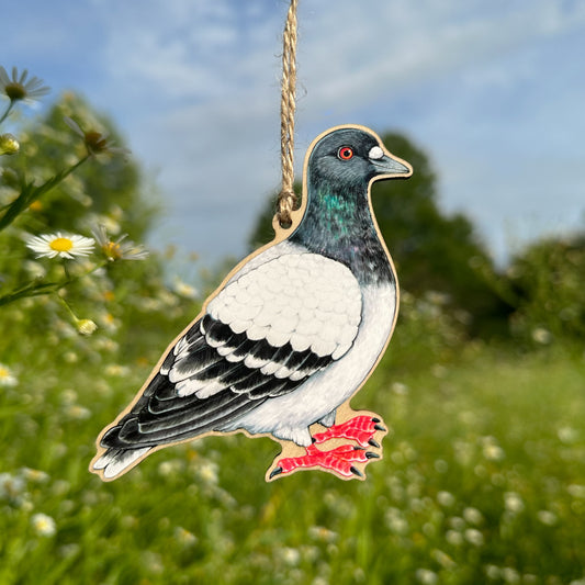 A wooden pigeon ornament with a hanging twine string, set against a blurred outdoor background of green grass, white flowers, and blue sky.