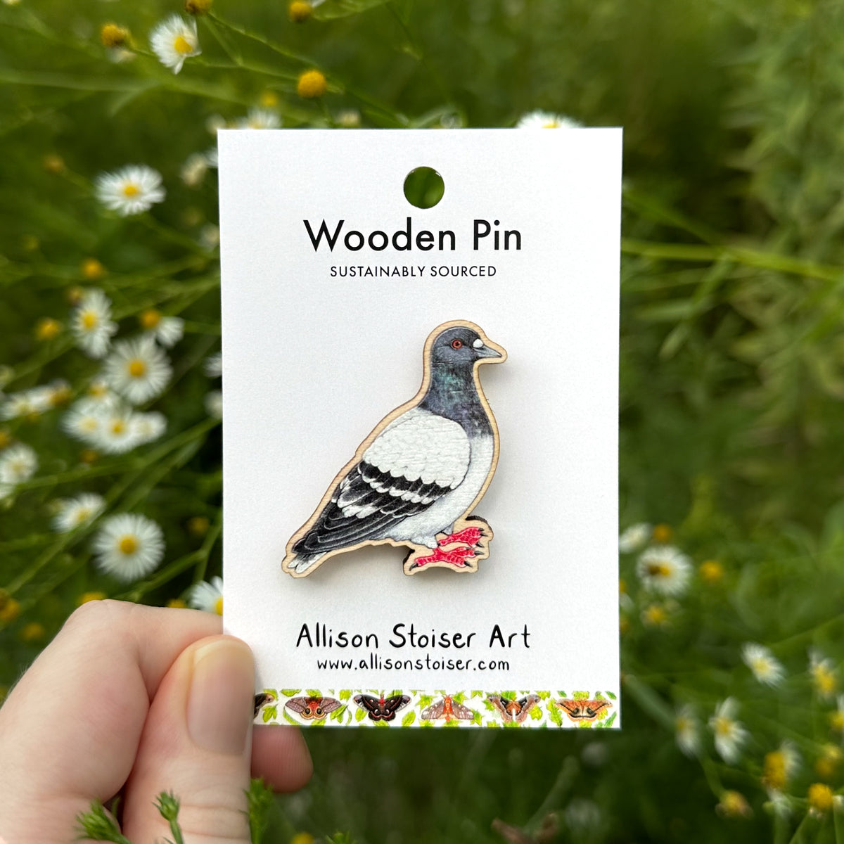 A wooden pigeon pin by Allison Stoiser Art is displayed on its white backing card, held by a hand against a blurred background of green foliage and asters.