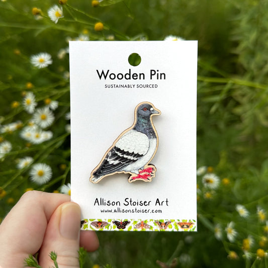 A wooden pigeon pin by Allison Stoiser Art is displayed on its white backing card, held by a hand against a blurred background of green foliage and asters.