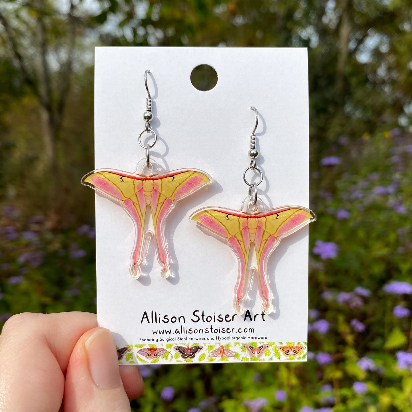 Pink spirit moth earrings on a card held by a hand with a natural background