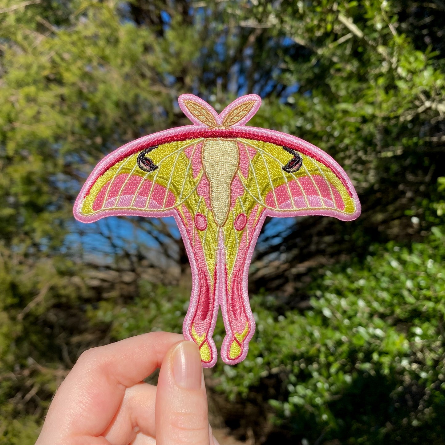 An embroidered patch depicting a pink spirit moth held against a blurred natural background.
