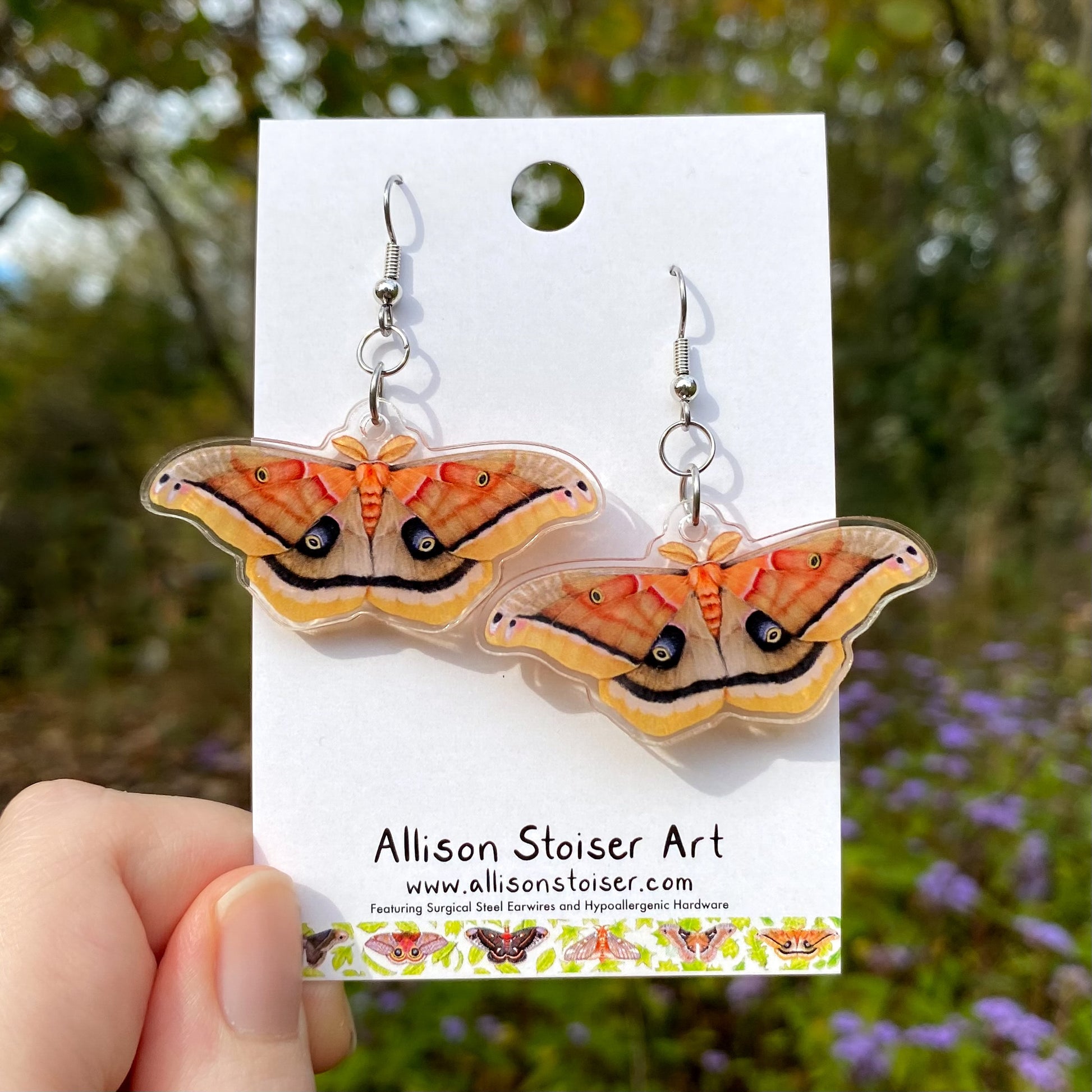 Polyphemus moth earrings on a card held by a hand with a natural background