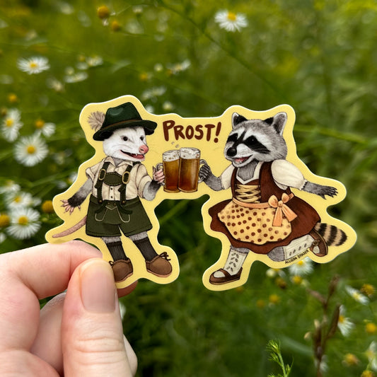 A hand holds a die-cut sticker featuring an illustrated opossum in lederhosen and raccoon with a dirndl with beer, with the word 'PROST!' between them, set against a blurred green wildflower background.