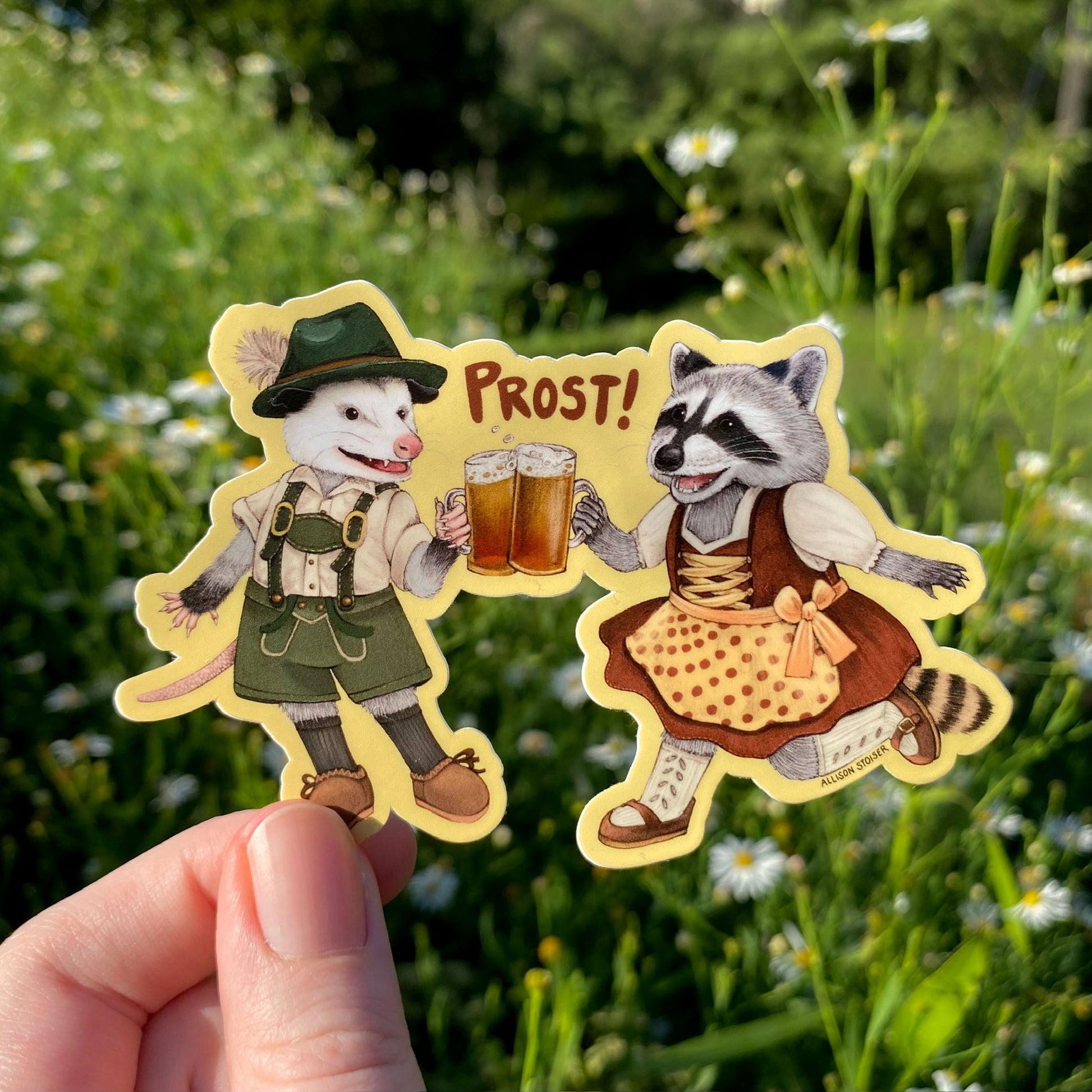 A sticker of an opossum and raccoon holding beer glasses with 'PROST!' text, held against a natural background.