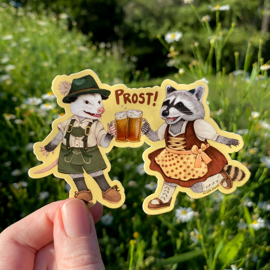 A sticker of an opossum and raccoon holding beer glasses with 'PROST!' text, held against a natural background.