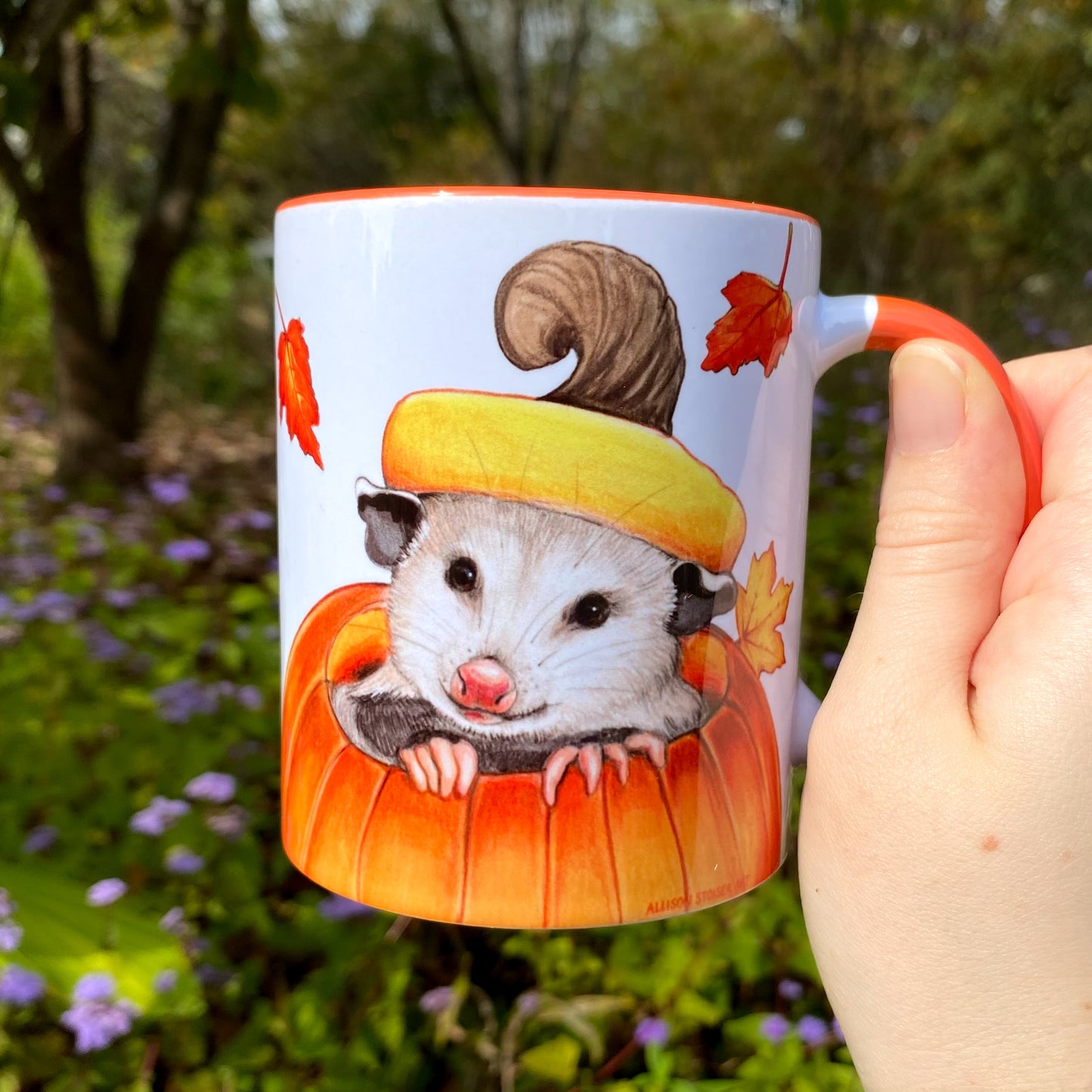 Mug with an illustration of an opossum in a pumpkin and autumn leaves held in front of a natural background.