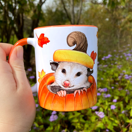 Mug with an opossum in a pumpkin held outdoors.