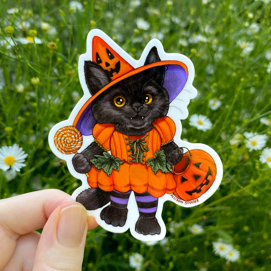 A die-cut sticker featuring a black cat dressed in an orange pumpkin witch costume, set against a blurred green grassy background with small white flowers.