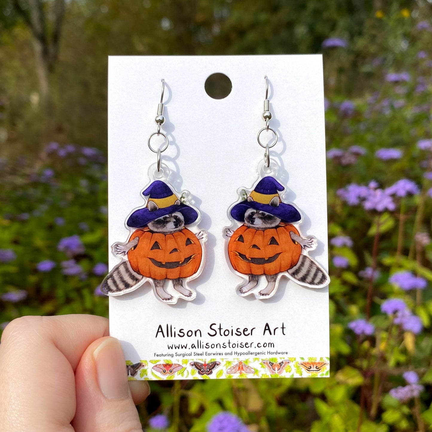 Earrings of a raccoon in a pumpkin costume on a card with a blurred natural background