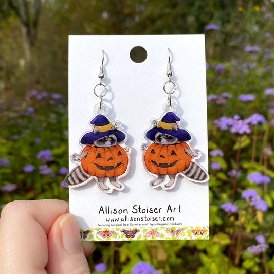 Earrings of a raccoon in a pumpkin costume on a card with a blurred natural background