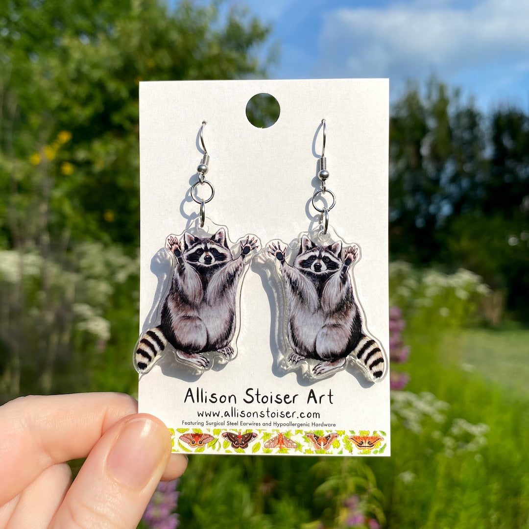 A hand holding a pair of acrylic earrings of raccoons with their hands raised in the air.