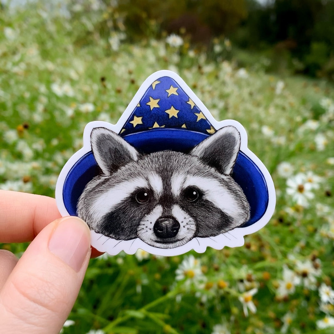 A hand holding a sticker of a raccoon wearing a blue wizard hat with gold stars.