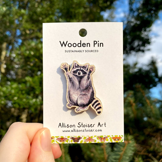 A hand holding a wooden pin with a raccoon design on it, sustainably sourced, with a rubber clutch on the back, displayed against a blurred background.