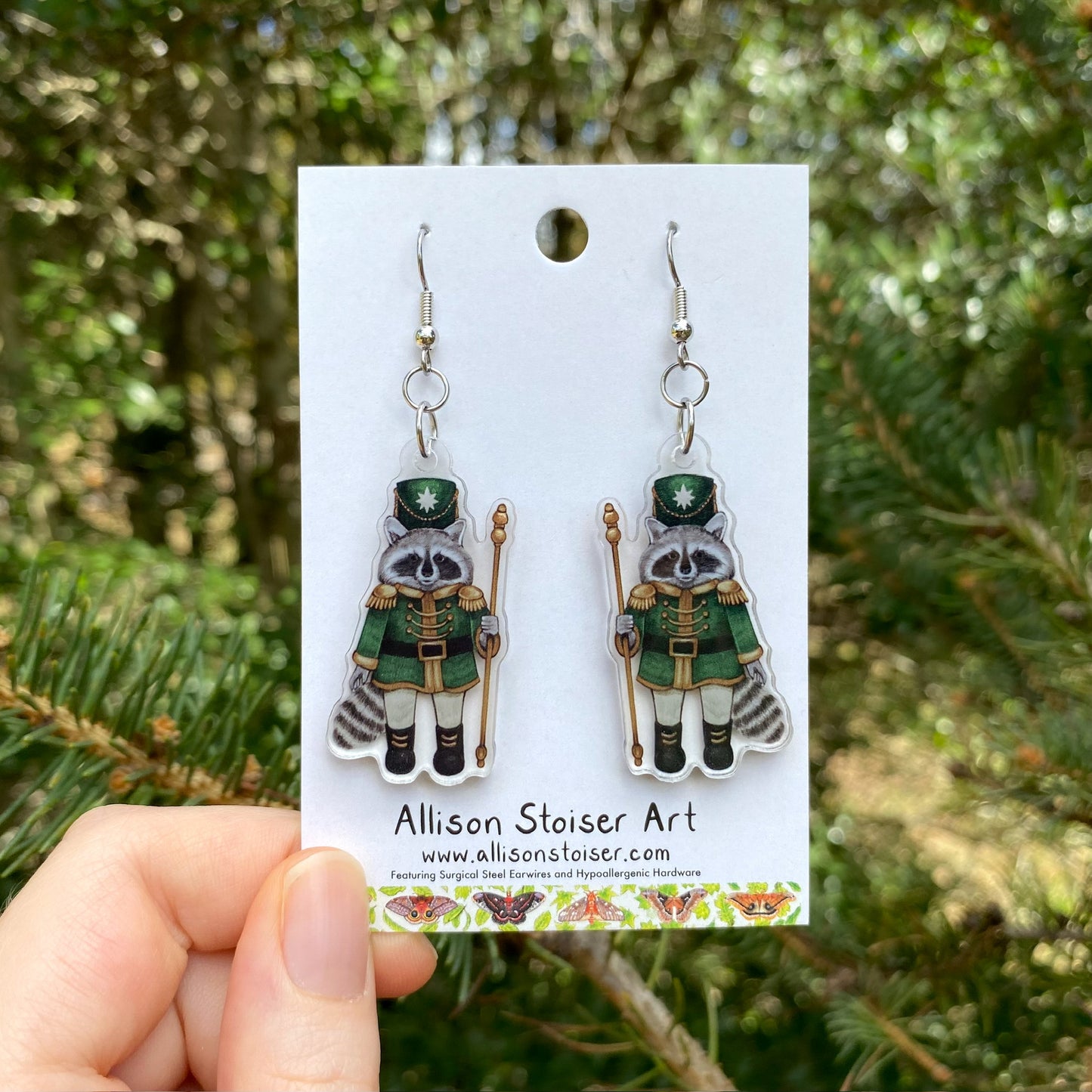 A hand holding a pair of raccoon nutcracker acrylic earrings