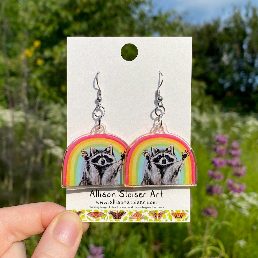 A hand holding a pair of acrylic earrings with raccoons in front of rainbows.