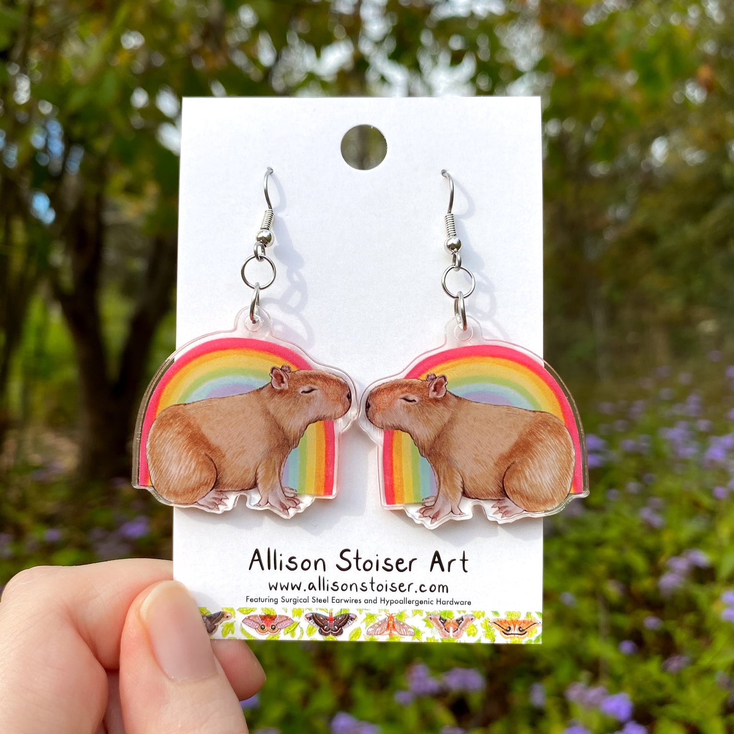 Capybara earrings with rainbow design on an Allison Stoiser Art branded card, held against a natural background.