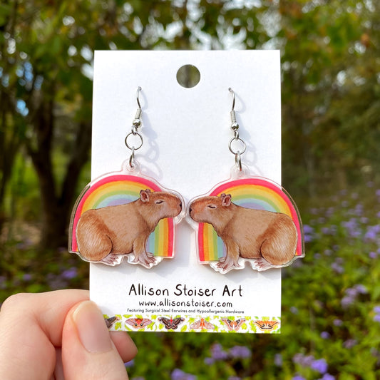 Capybara earrings with rainbow design on an Allison Stoiser Art branded card, held against a natural background.