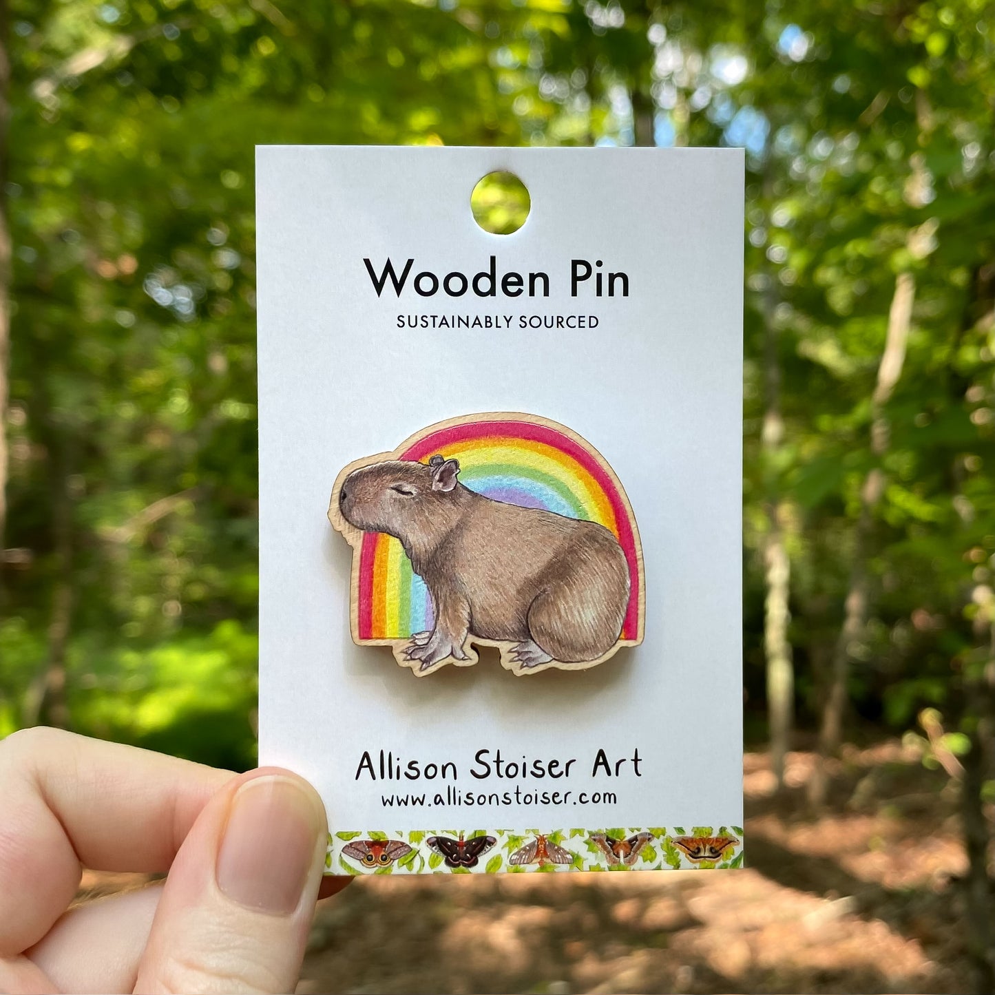 Wooden pin with capybara in front of a rainbow on a card with 'Allison Stoiser Art' branding, held in front of a forest background.