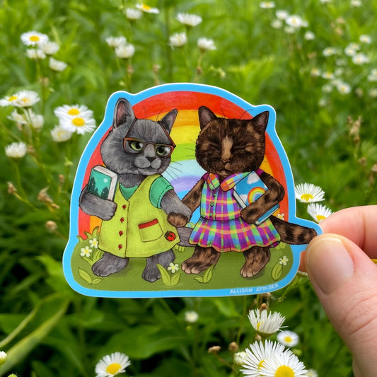 Sticker of two cats holding hands with a rainbow background, held in front of greenery and flowers.