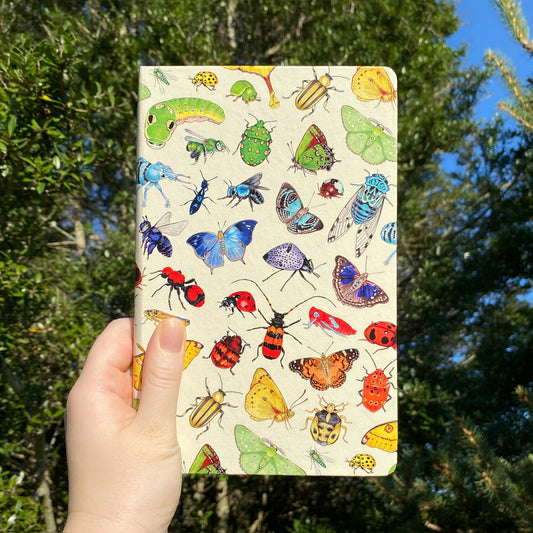 A hand holding an A5 leatherette journal with a colorful rainbow insects print, in front of a tree.