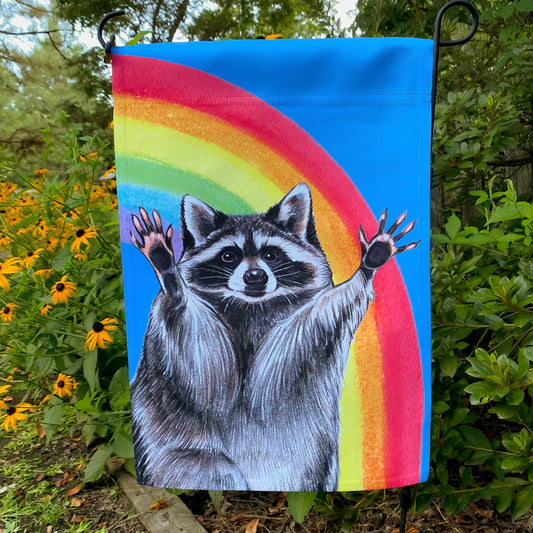 A garden flag of a raccoon with its arms raised in front of a rainbow.