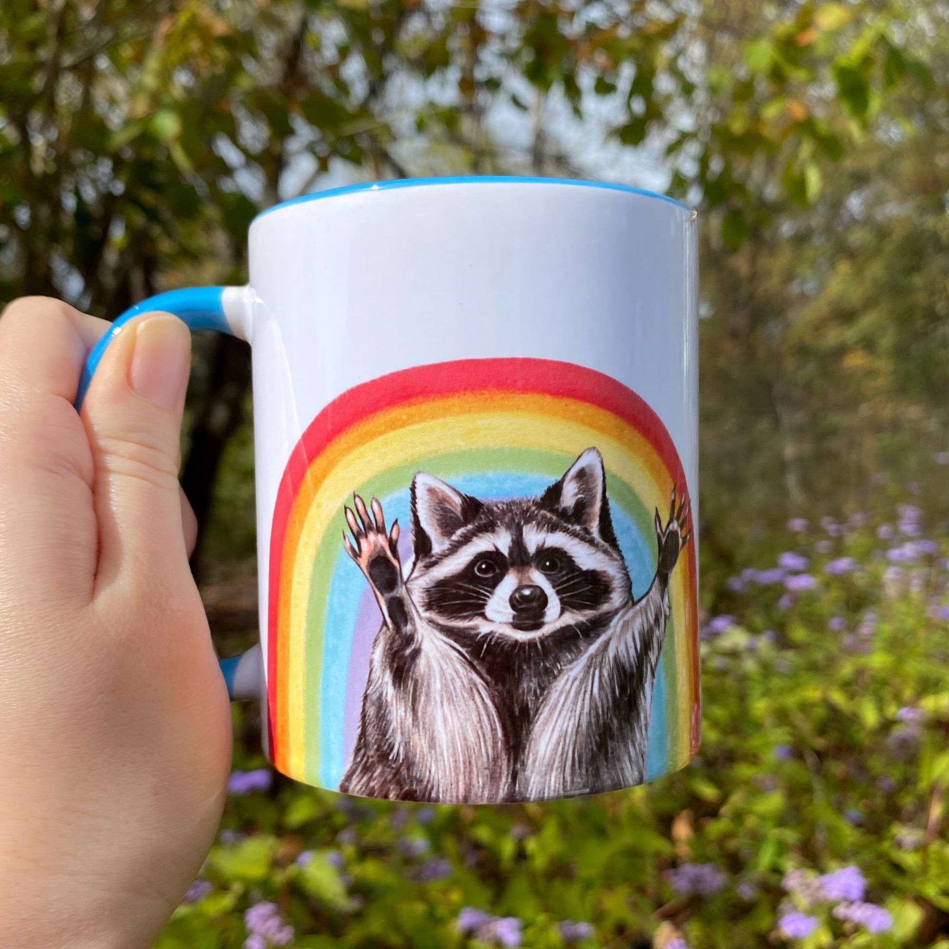 Mug with a raccoon illustration in front of a rainbow held by a hand outdoors.