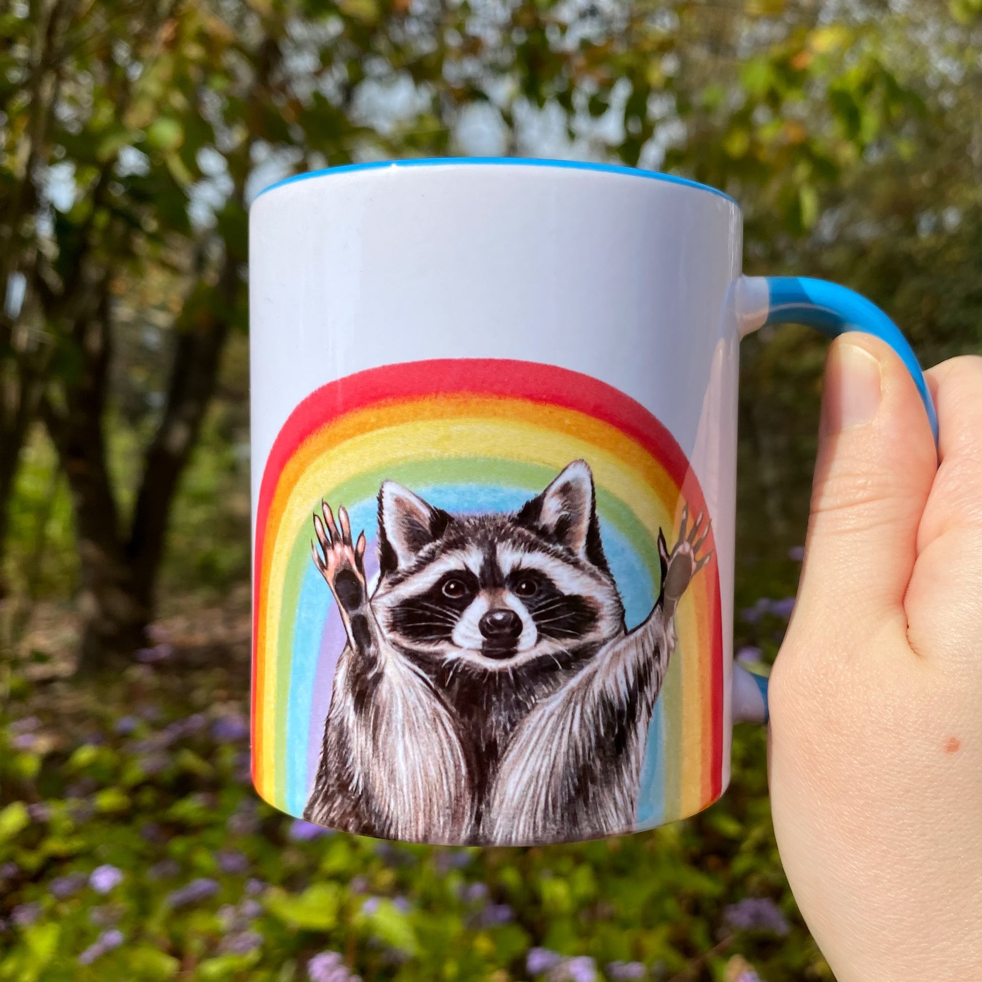 Mug with a raccoon and rainbow illustration held in front of a natural background