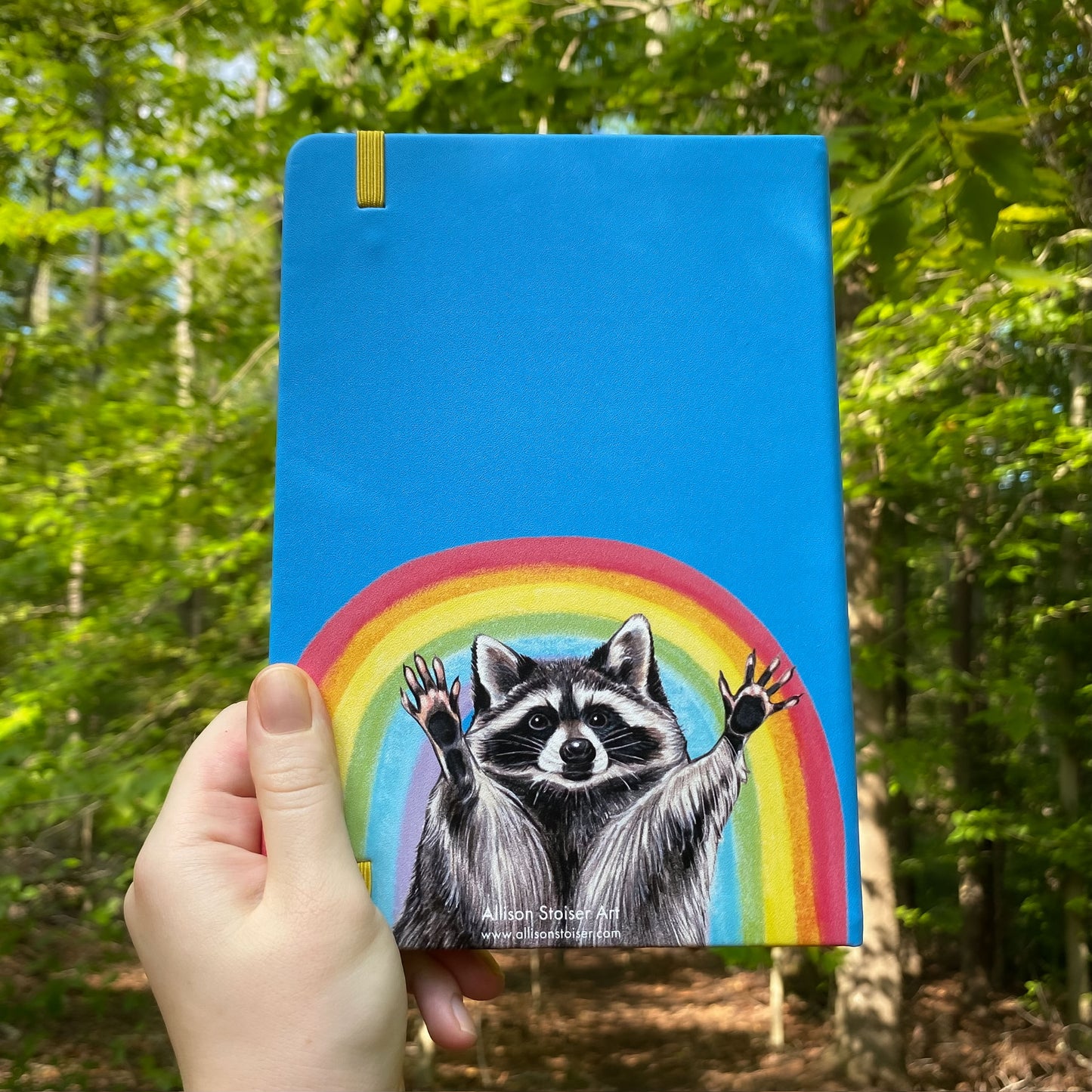 Notebook with a raccoon and rainbow held in front of a forest background
