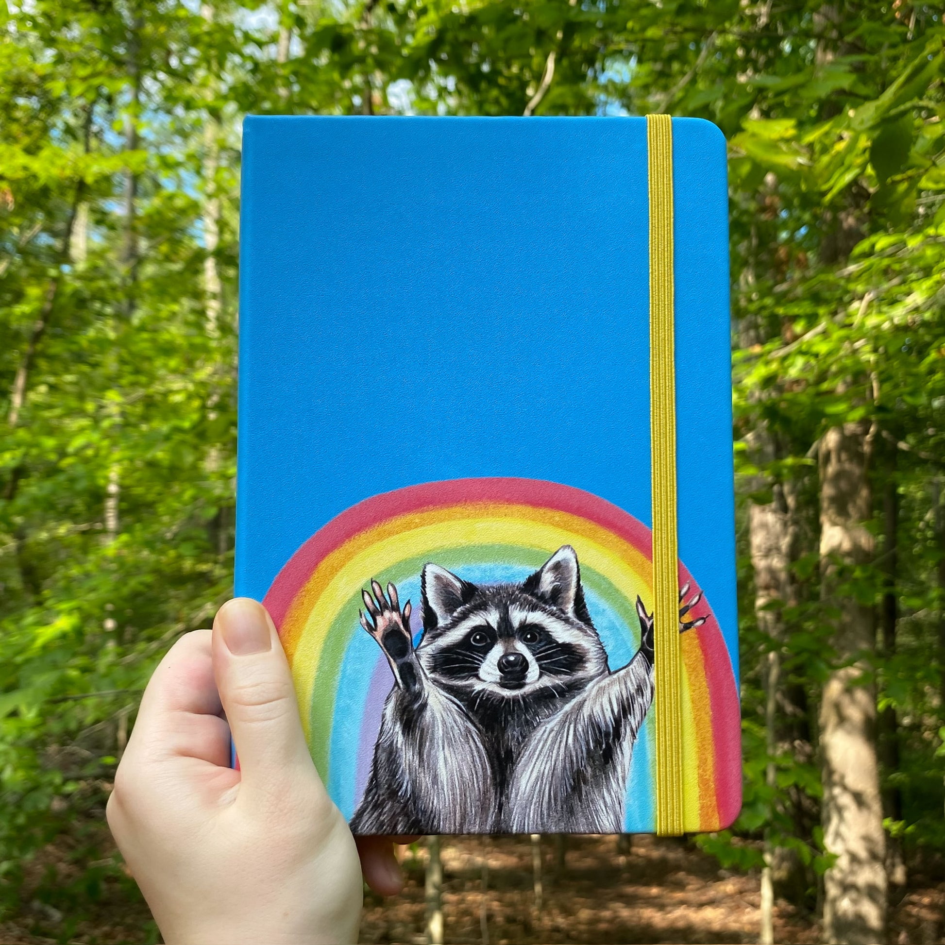 Blue notebook with a raccoon and rainbow held in front of a forest