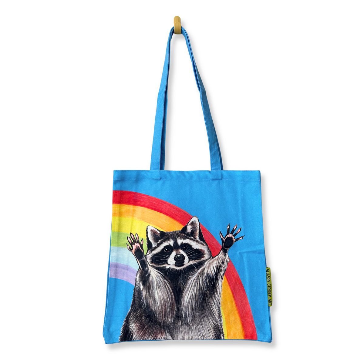 A bright blue canvas tote bag with long handles, featuring a printed illustration of a raccoon with raised paws in front of a rainbow. The bag is hanging from a small wooden hook against a plain white background.