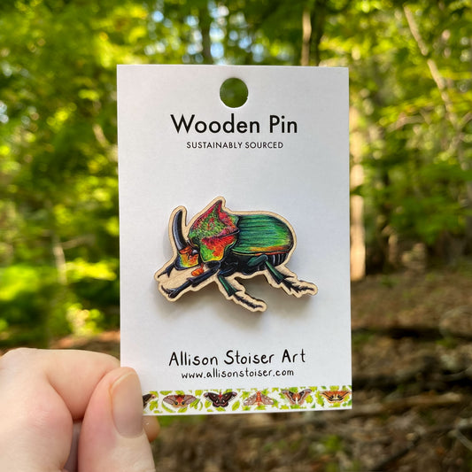 Wooden pin of a rainbow scarab beetle on a card with 'Allison Stoiser Art' branding, held against a natural background.