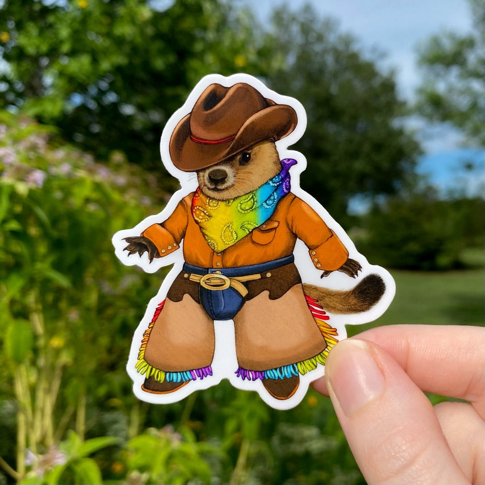 A hand holding a sticker of a groundhog dressed up in a rainbow cowboy outfit.