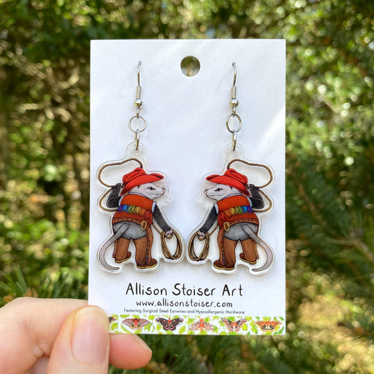 A hand holding a pair of cowboy opossum acrylic earrings