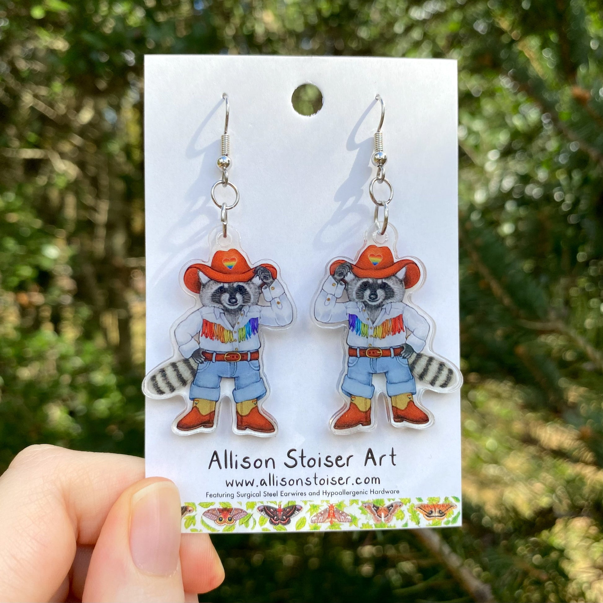 A hand holding a pair of cowboy raccoon acrylic earrings