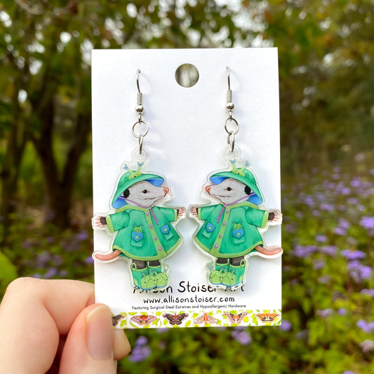 Pair of earrings with opossums in green raincoats on a blurred nature background