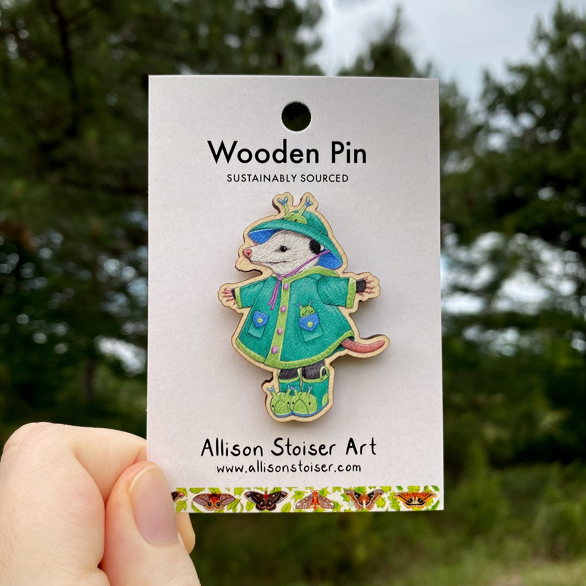Wooden pin of an opossum in a green raincoat on a card with 'Allison Stoiser Art' branding, against a blurred natural background.