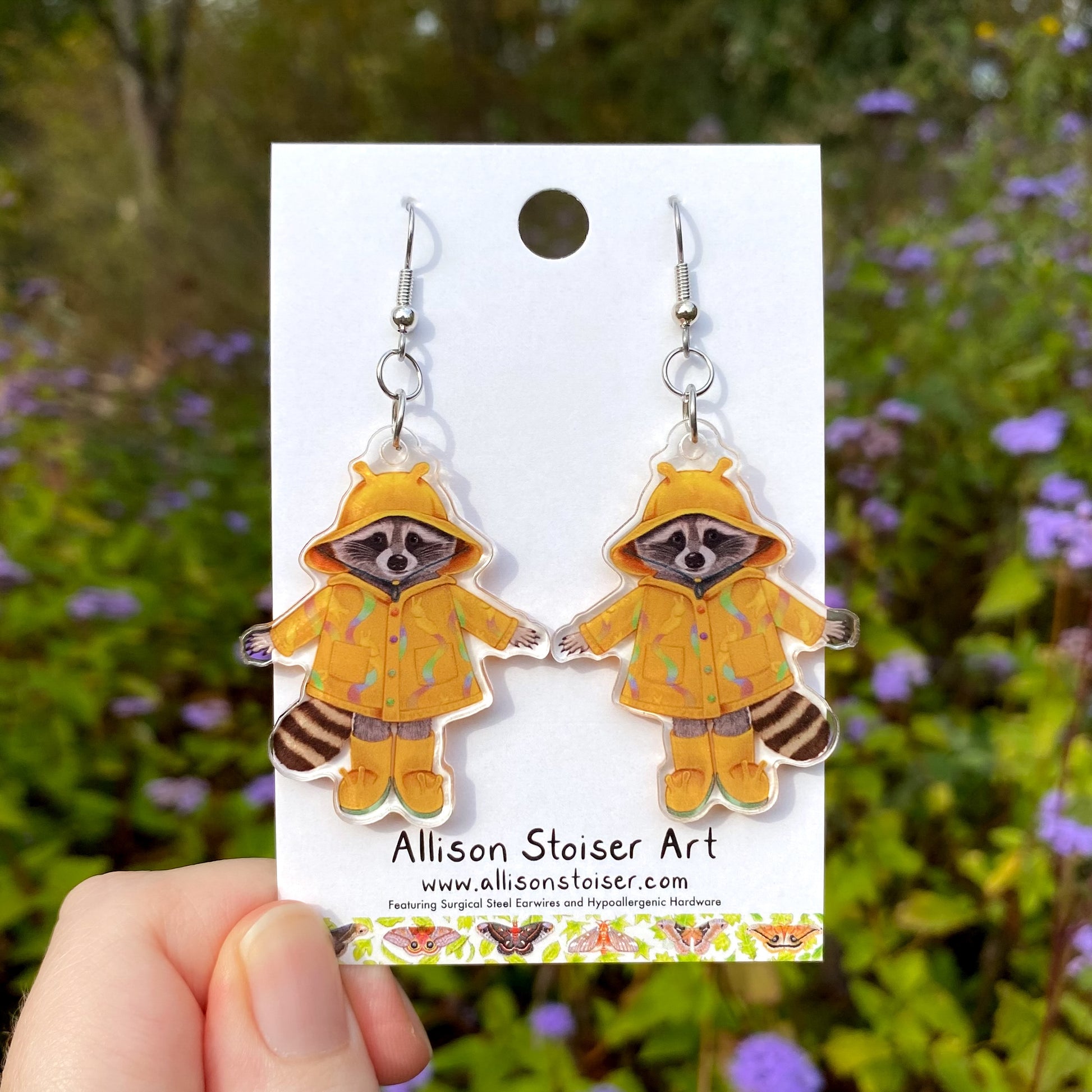 Raccoon earrings in a raincoat on a card with a blurred floral background