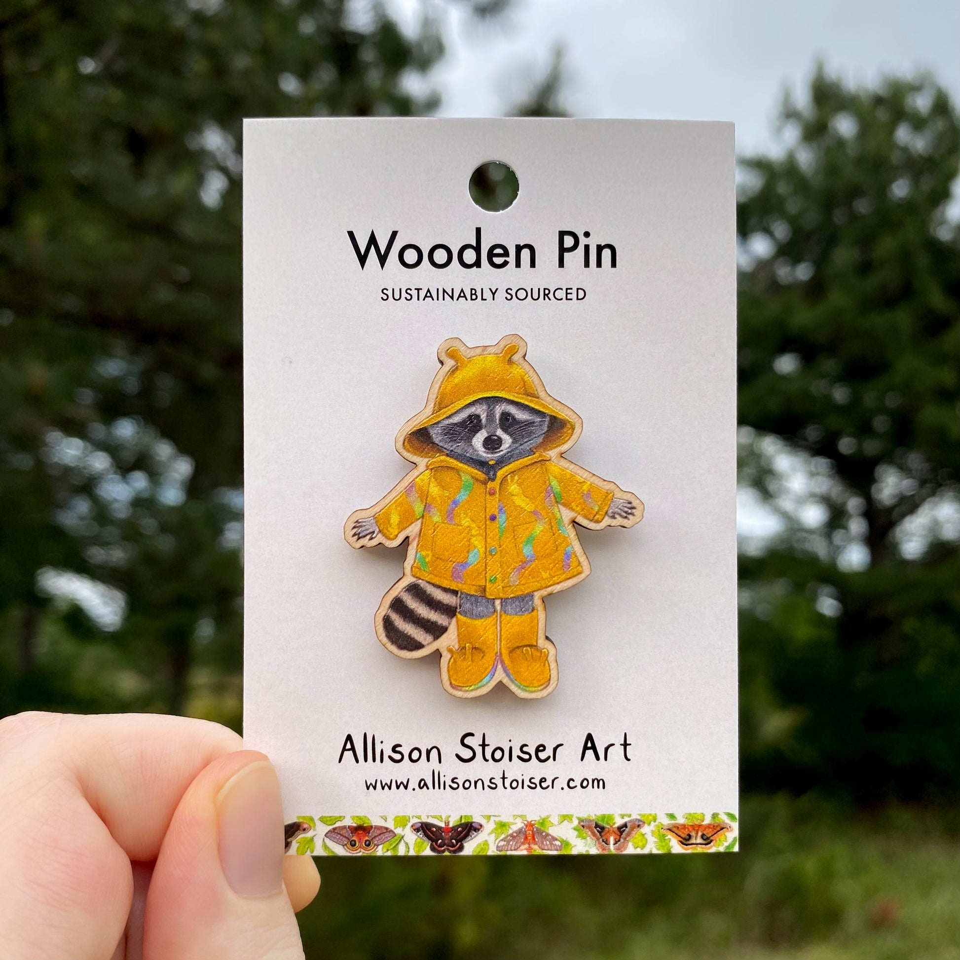 Wooden pin of a raccoon in a yellow raincoat on a card with 'Allison Stoiser Art' branding, against a blurred natural background.