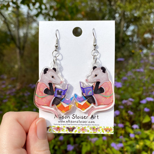Pair of earrings with reading opossums on a card held in front of a natural background.