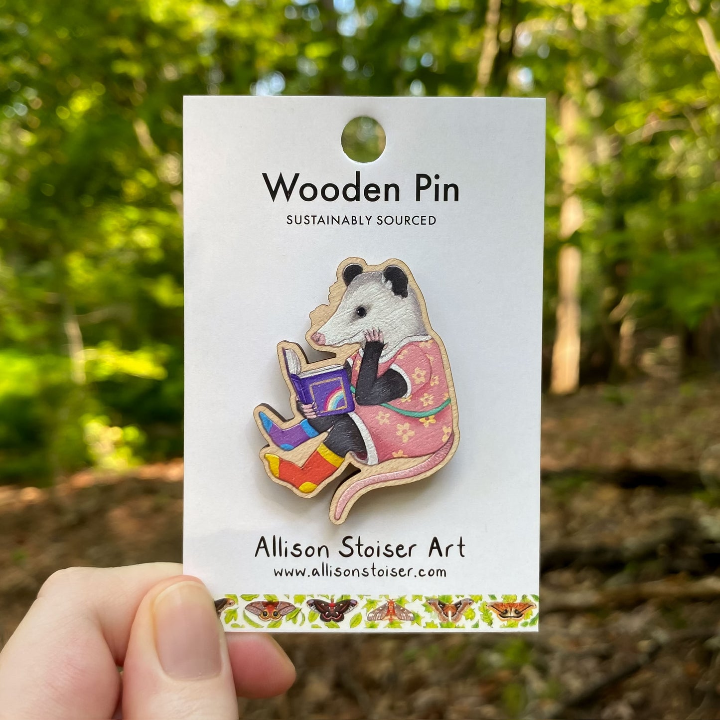 Wooden pin of an opossum reading a book on a card with 'Allison Stoiser Art' branding, held against a natural background.