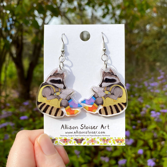Reading raccoon earrings on a card held in front of a natural background