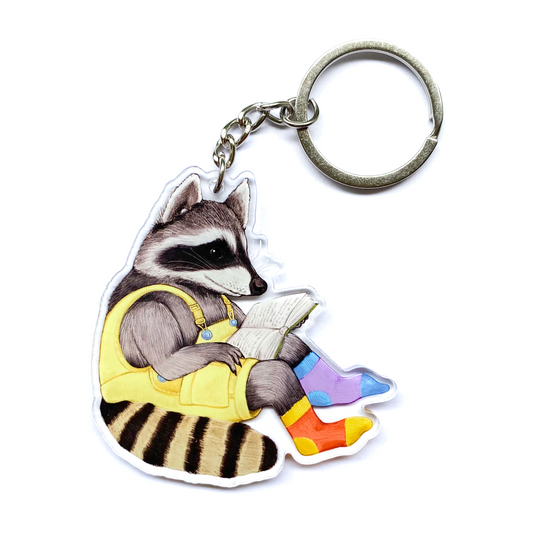 Keychain of a raccoon wearing overalls and reading a book on a white background