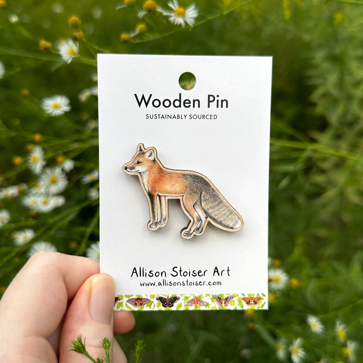 A wooden fox pin by Allison Stoiser Art, mounted on a white backing card and held by a hand against a blurred background of green foliage and asters.