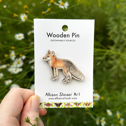 A wooden fox pin by Allison Stoiser Art, mounted on a white backing card and held by a hand against a blurred background of green foliage and asters.