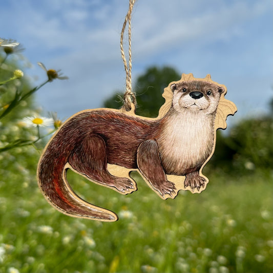 A wooden otter ornament with twine thread, shown outdoors against a blurred green field and blue sky background.