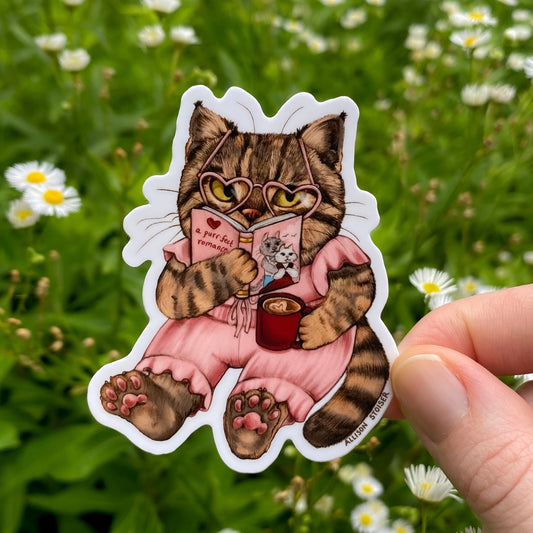 Sticker of a cat reading a book with heart-shaped glasses held in front of a natural background with flowers.