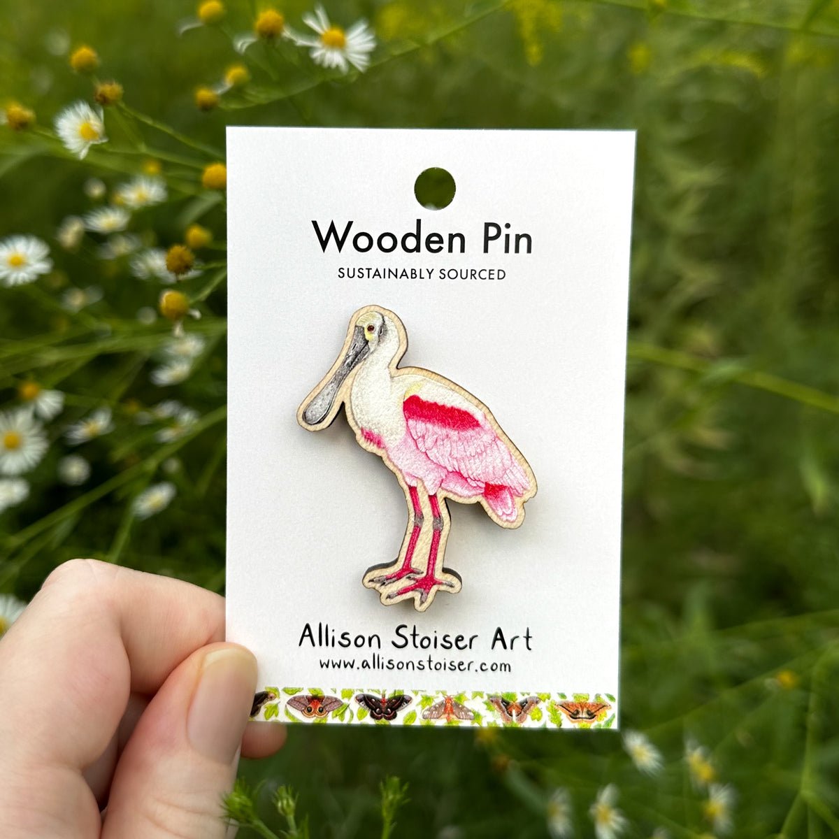 A wooden pin of a roseate spoonbill on a white backing card held by a hand against a blurred green outdoor background of asters.