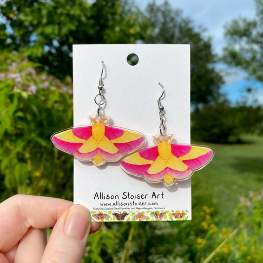 A hand holding a pair of acrylic rosy maple moth earrings.