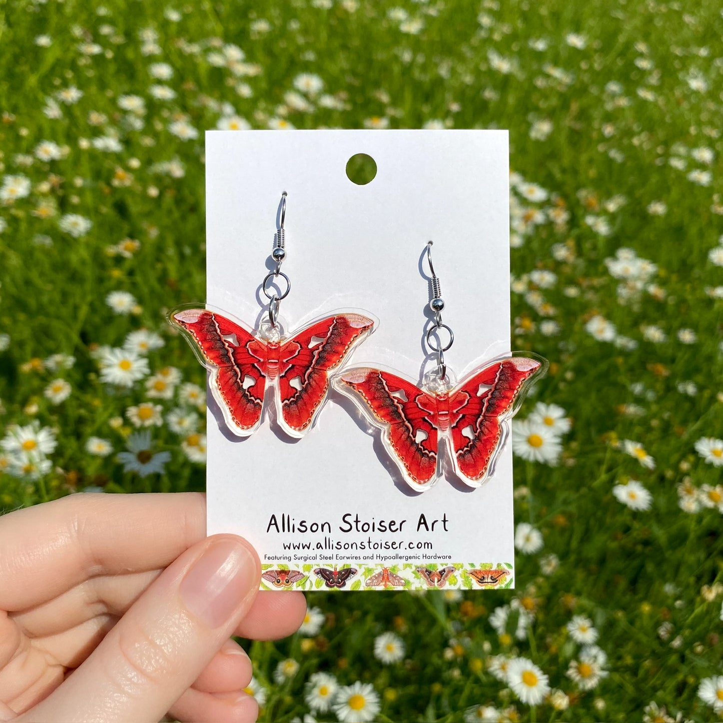 A hand holding a pair of clear acrylic earrings of red Rothschildia moths.