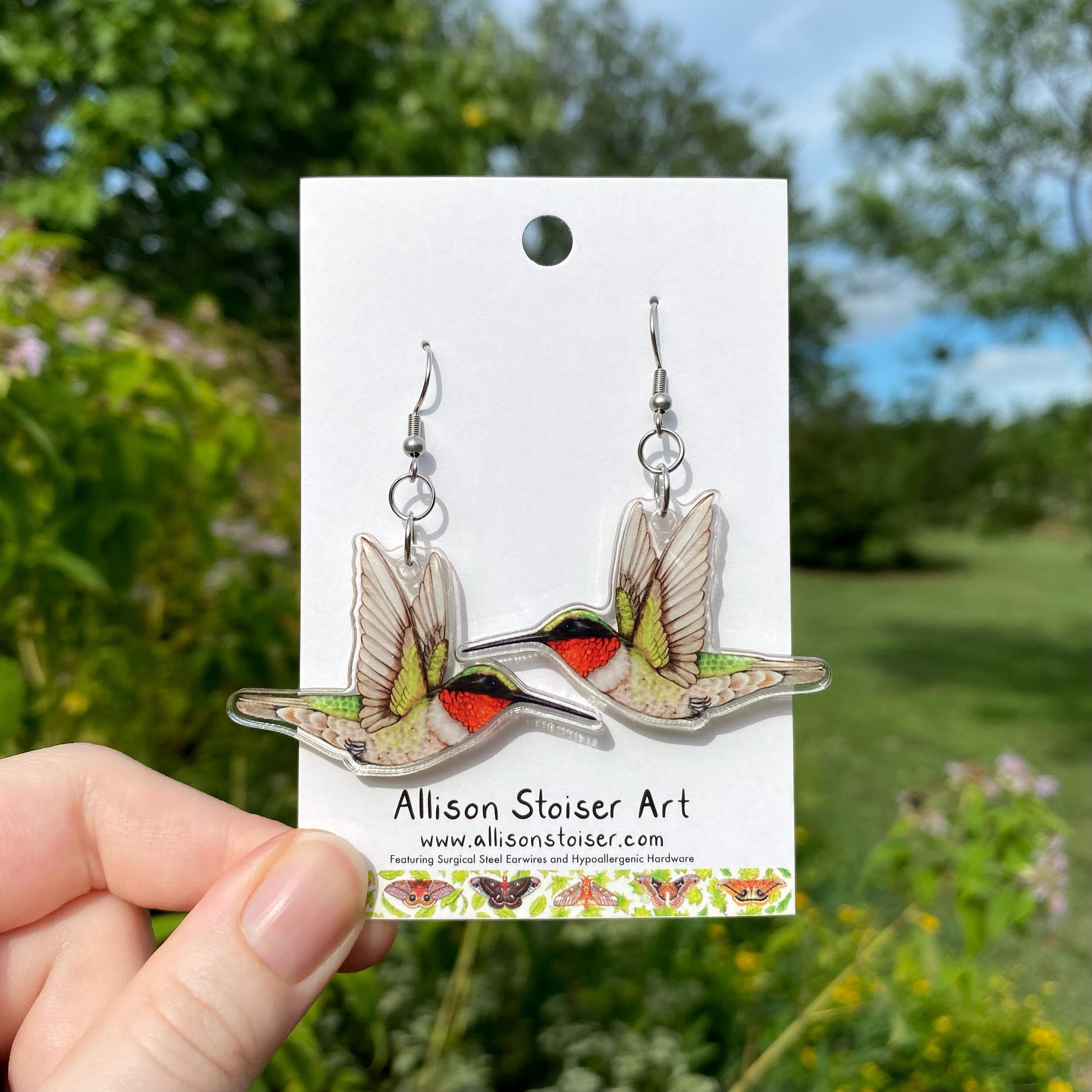 A hand holding a pair of acrylic ruby-throated hummingbird earrings.